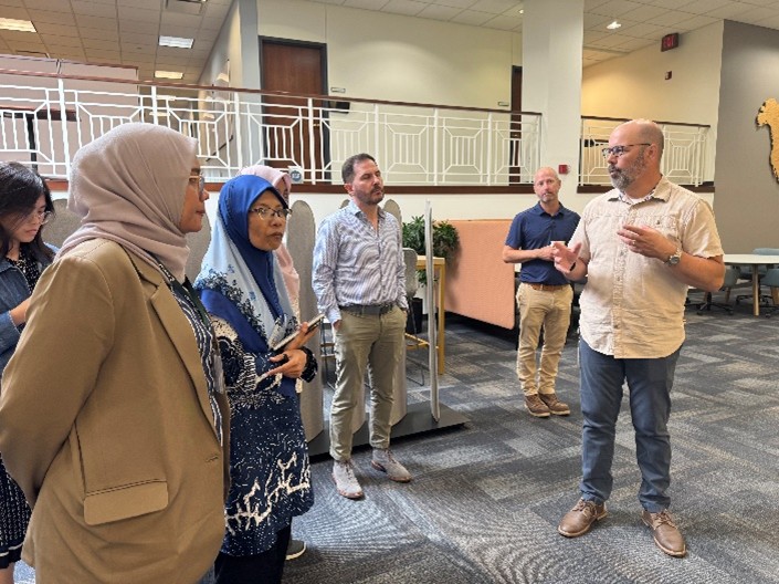 A group of participants stands in a building lobby while a man in the foreground speaks to them, gesturing with his hands. The group appears to be listening during a guided tour or briefing. The space includes a railing on the upper level, office doors and seating areas in the background.
